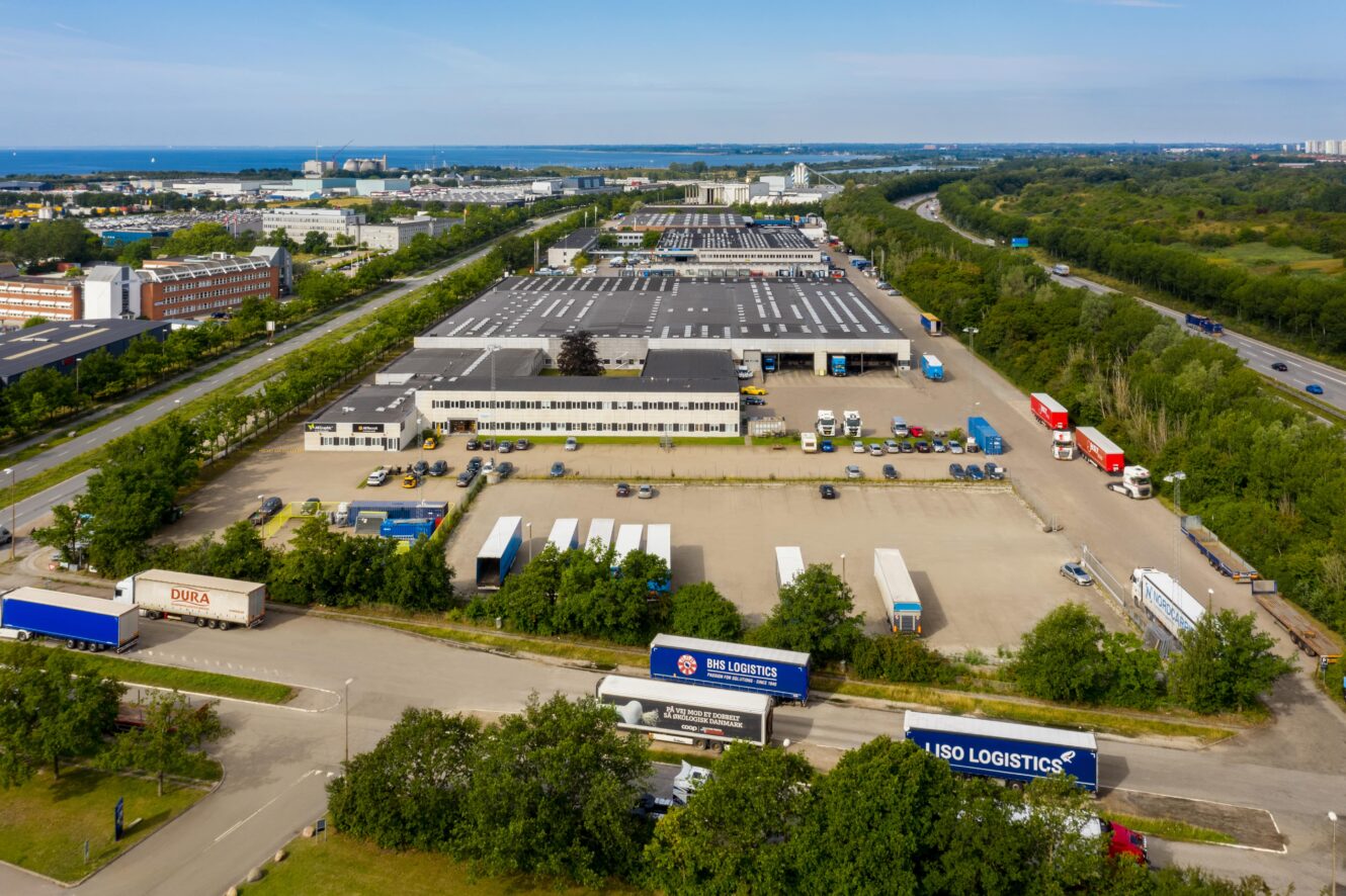 Luftfoto af et stort logistik- og distributionscenter med lagerbygninger, mange lastbiler og trailere parkeret på læssepladserne, omgivet af grønne områder og veje.