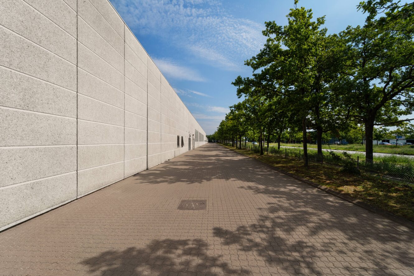 En lang, blank betonvæg løber parallelt med en gangsti med grønne træer under en blå himmel med spredte skyer.
