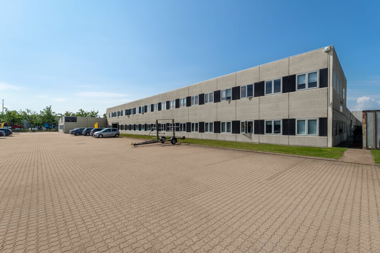 Rektangulær kontorbygning i to etager med mange vinduer, omgivet af en stor asfalteret parkeringsplads med flere parkerede biler og klar blå himmel.