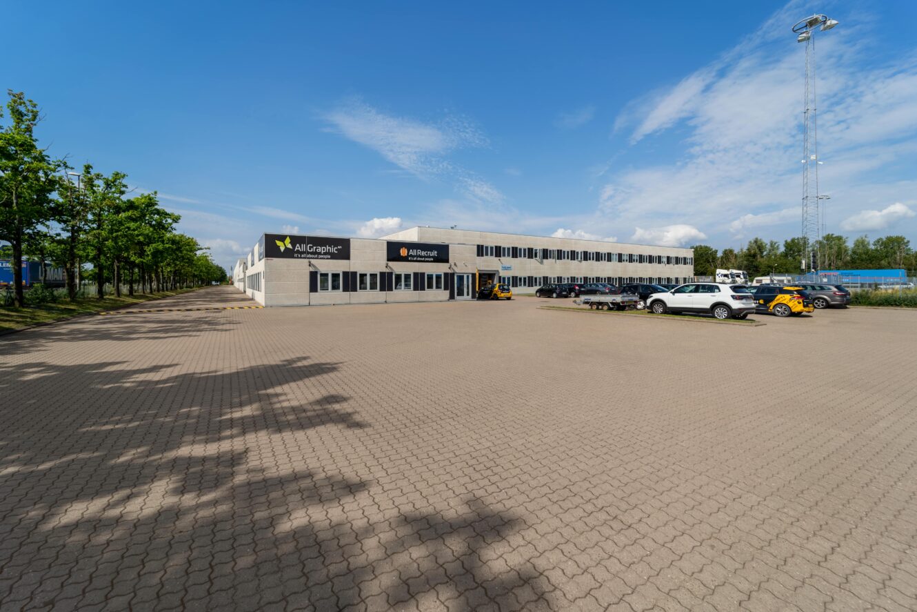 Stor industribygning med flere køretøjer parkeret foran på en bred asfalteret grund, træer langs siden og klar blå himmel over.