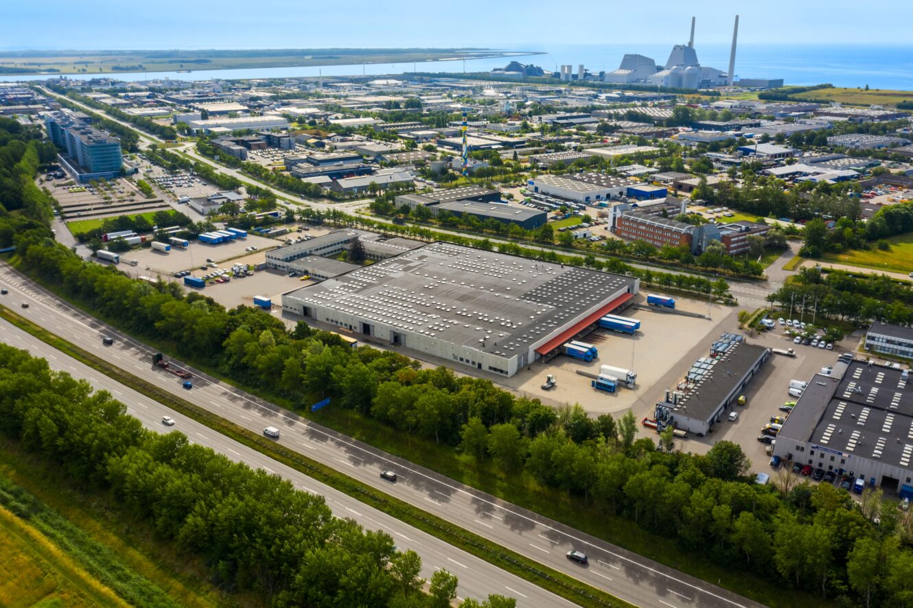 Luftfoto af et industriområde med lagerbygninger, fabrikker og parkeringspladser ved siden af en motorvej med vand- og kraftværksstrukturer i baggrunden.