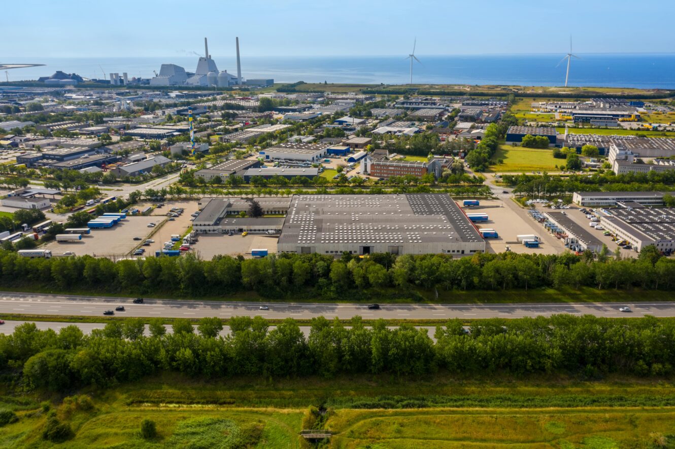 Luftfoto af et industriområde med lagerbygninger, fabrikker, parkerede lastbiler, vindmøller og en motorvej i nærheden af en kystlinje under en klar himmel.
