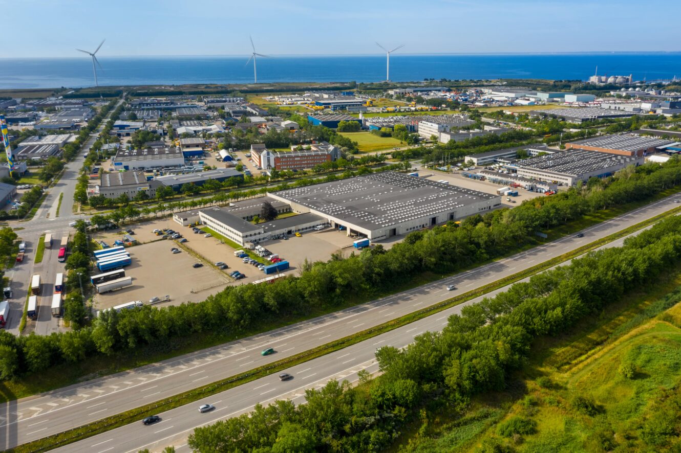 Luftfoto af et industriområde med lagerbygninger, lastbiler, vindmøller og en nærliggende motorvej tæt på kysten.