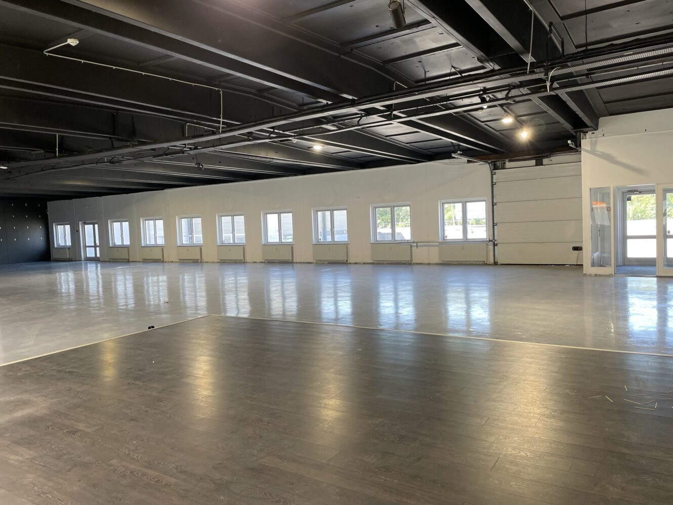 Stort, tomt rum med mørkt loft, store vinduer langs den ene væg og sektioner med lyst og mørkt gulv.