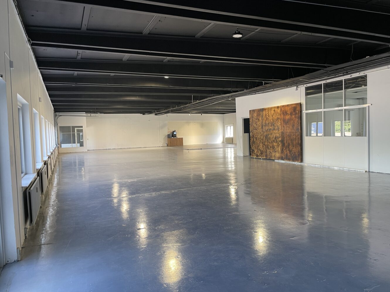 Stort tomt rum i industristil med poleret betongulv, hvide vægge, synlige sorte loftsbjælker og vinduer langs den ene side; en træskillevæg er synlig i baggrunden.
