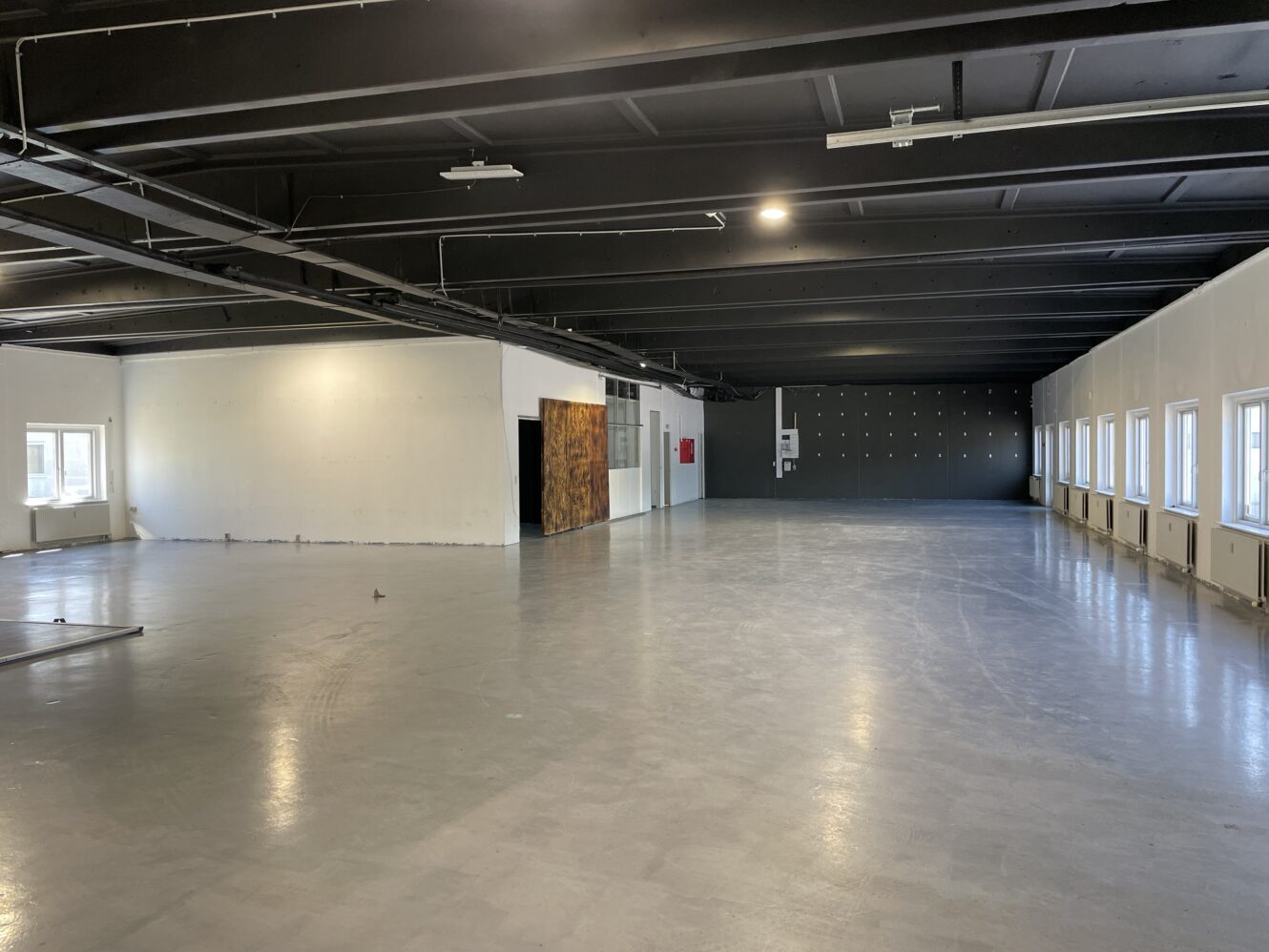 Stort tomt rum i industriel stil med polerede betongulve, hvide vægge, synligt loft og flere vinduer langs den ene side, der giver naturligt lys.