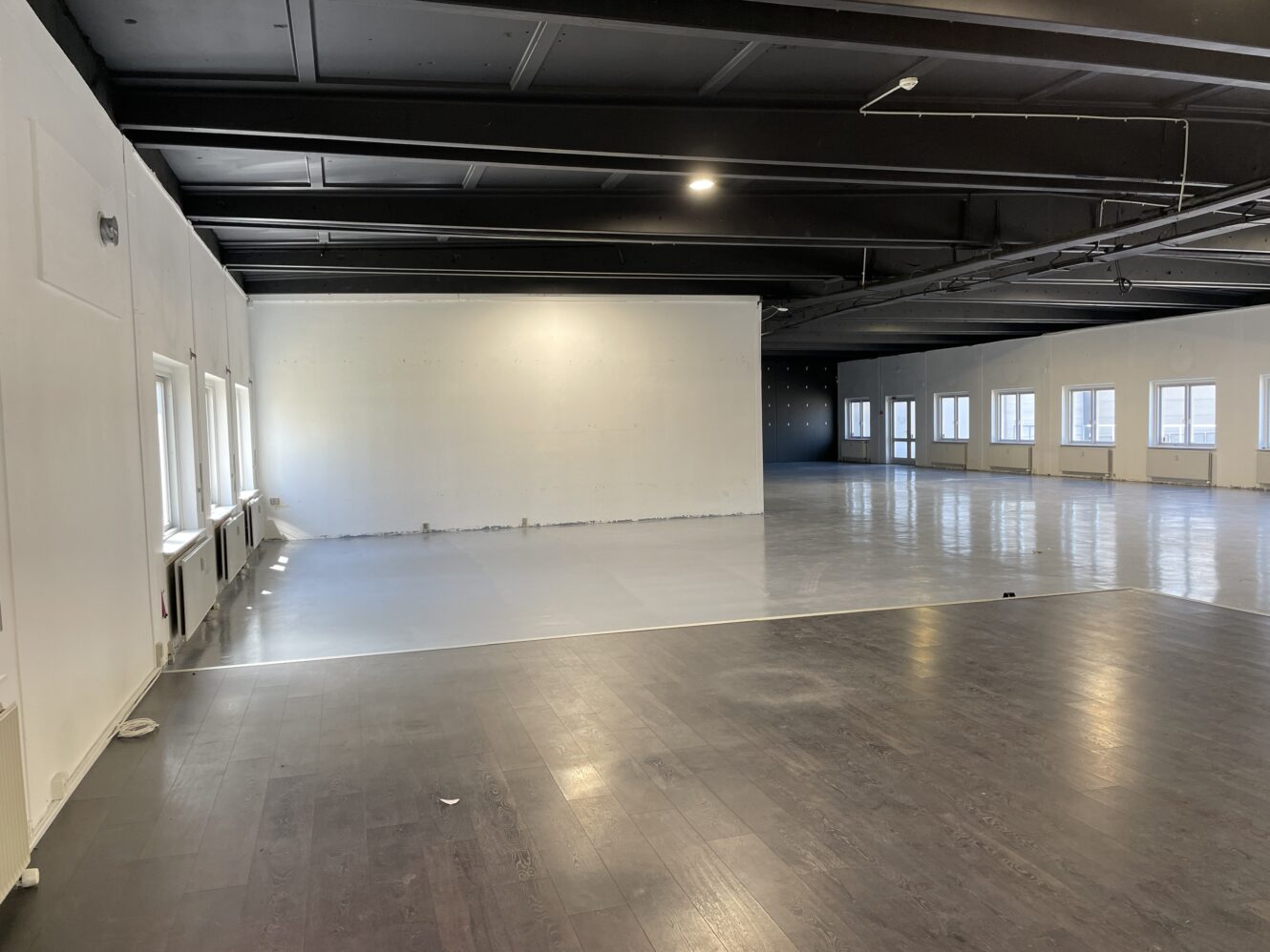 Stort, tomt rum med mørke og lyse gulve, hvide vægge, flere vinduer og synlige sorte loftsbjælker.