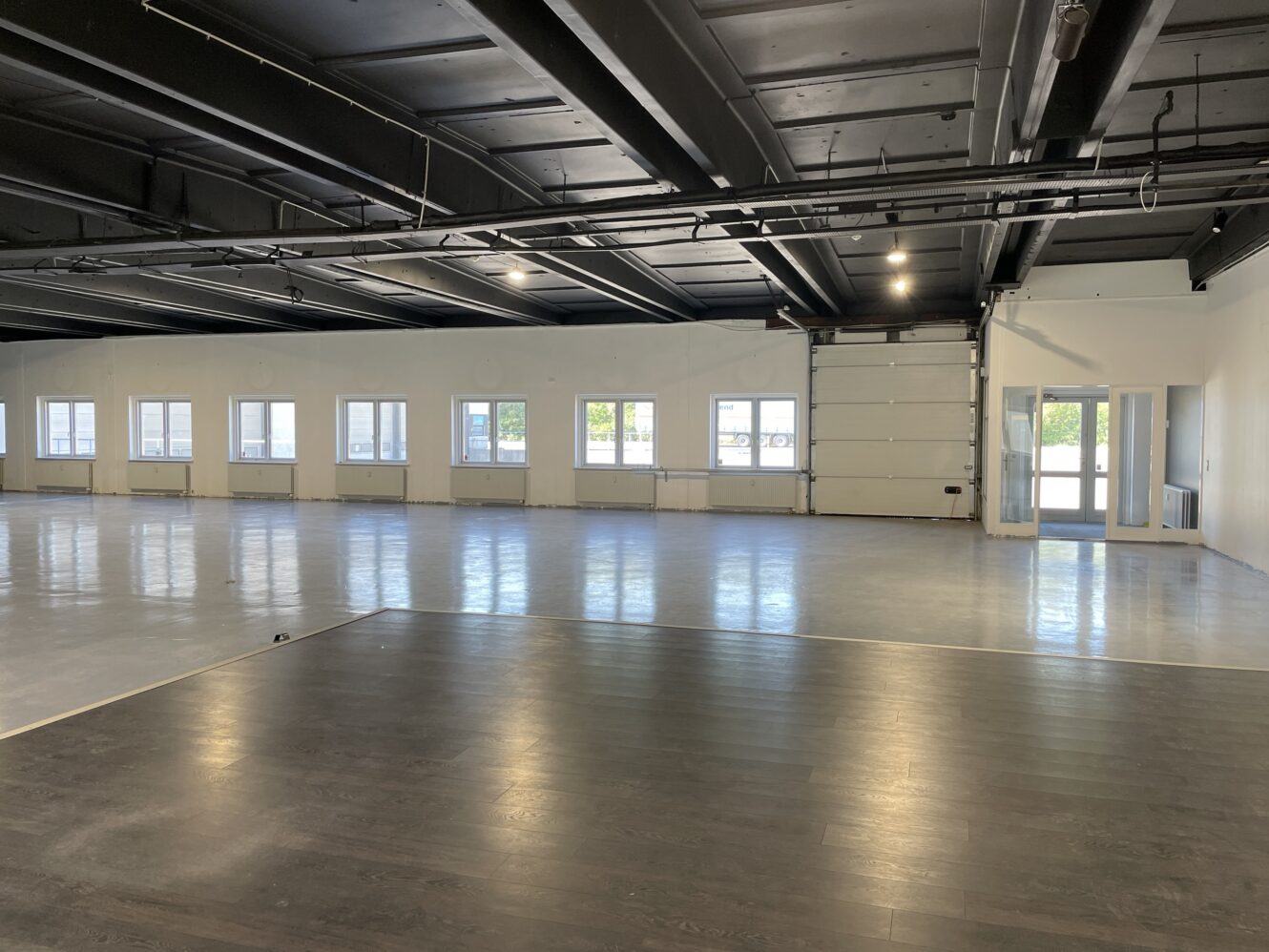 Stort, tomt industrielt rum med hvide vægge, flere vinduer, en glasdør og en kombination af mørkt træ og betongulv.