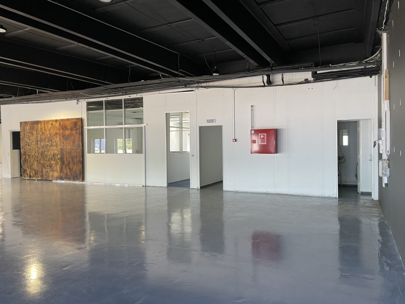 Et stort, tomt industrilokale med polerede betongulve, hvide vægge, indvendige vinduer, åbne døråbninger og et rødt brandslangeskab monteret på væggen.