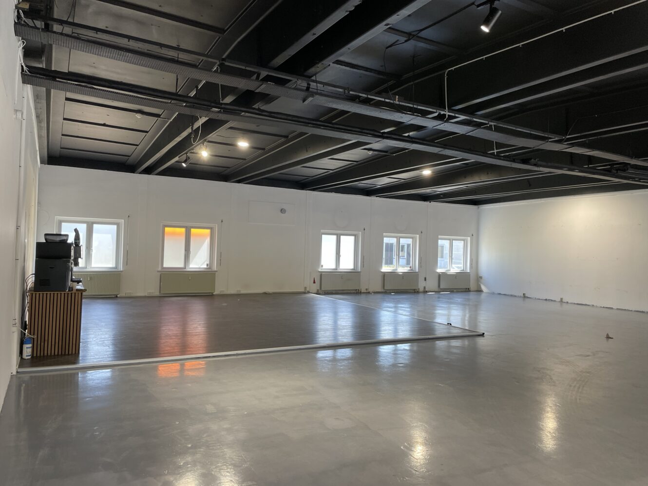 Rummeligt, tomt industrilokale med gråt gulv, hvide vægge, højt sort loft og flere vinduer langs den ene side, der lukker naturligt lys ind.