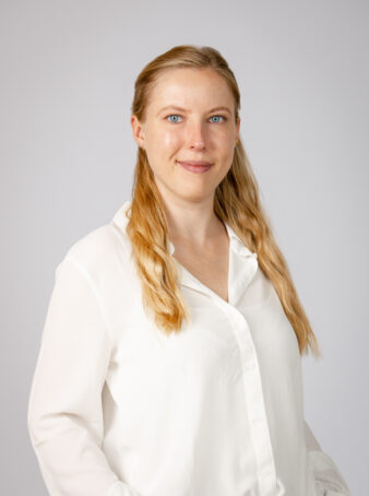 En kvinde med langt blondt hår og blå øjne, iført en hvid skjorte med knapper, står på en almindelig lysegrå baggrund.