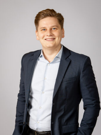 En ung mand i marineblå habitjakke og lyseblå skjorte står på en almindelig lysegrå baggrund og smiler til kameraet.