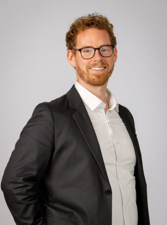 En mand med krøllet hår, briller og skæg er iført en mørk habitjakke over en hvid skjorte og står på en almindelig lys baggrund.