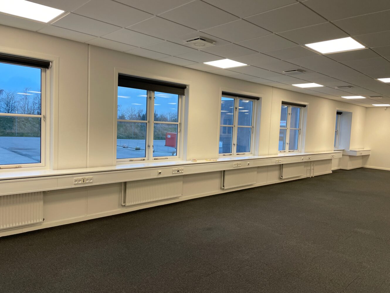 Stort, tomt kontorlokale med mørkt gulvtæppe, hvide vægge, loftslamper og flere vinduer langs den ene side, der lukker dagslys ind.