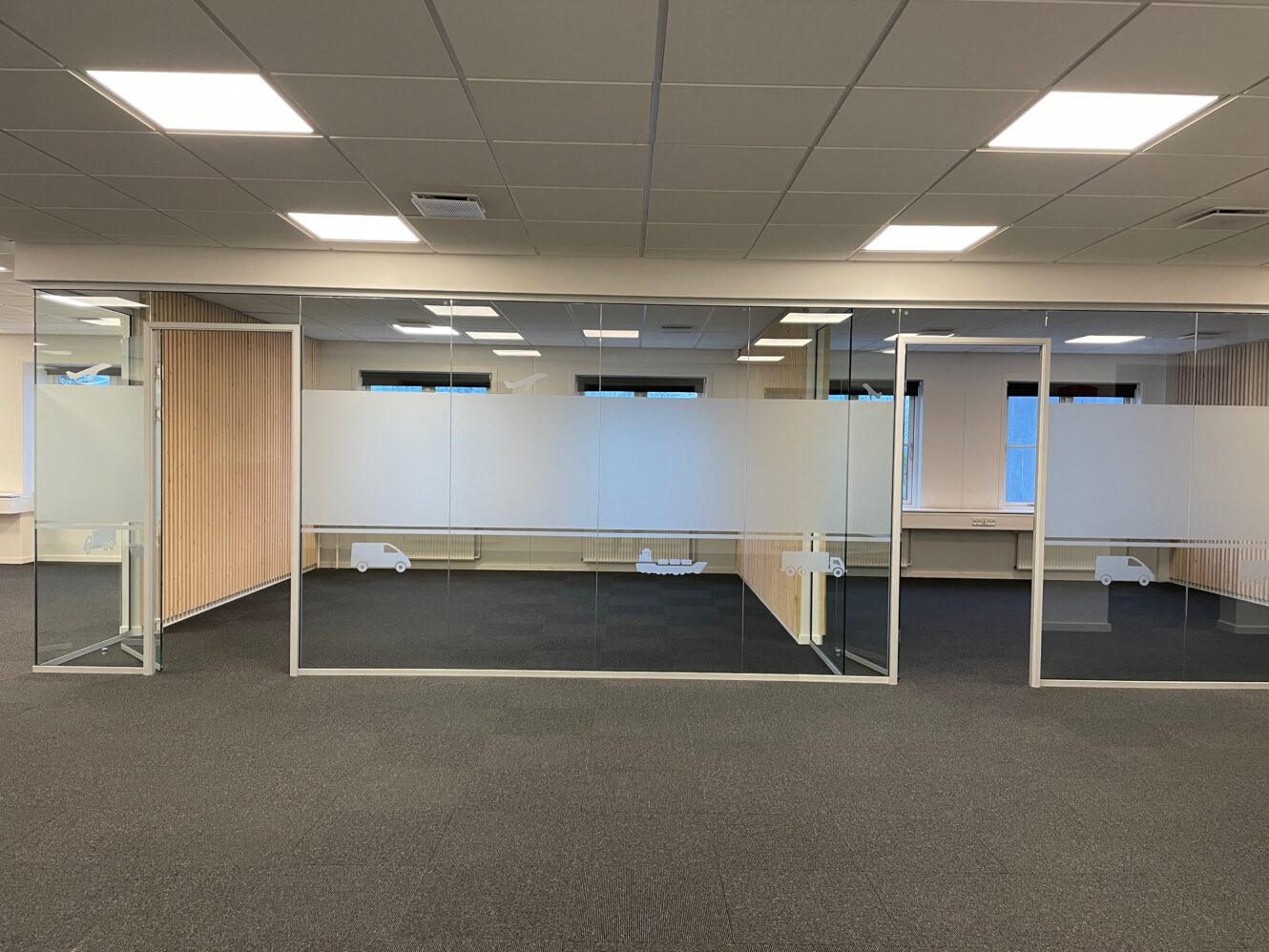 Moderne kontorlokaler med glasvægge i mødelokalerne, matterede afskærmningspaneler og minimalistisk indretning. Lokalet er tomt og godt oplyst med loftslamper.