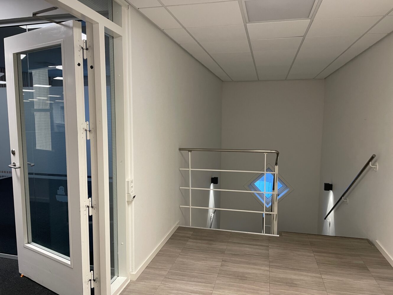 En gang med lyst klinkegulv, en åben dør til venstre og et rækværk med udsigt over en trappe og et lille diamantformet vindue.