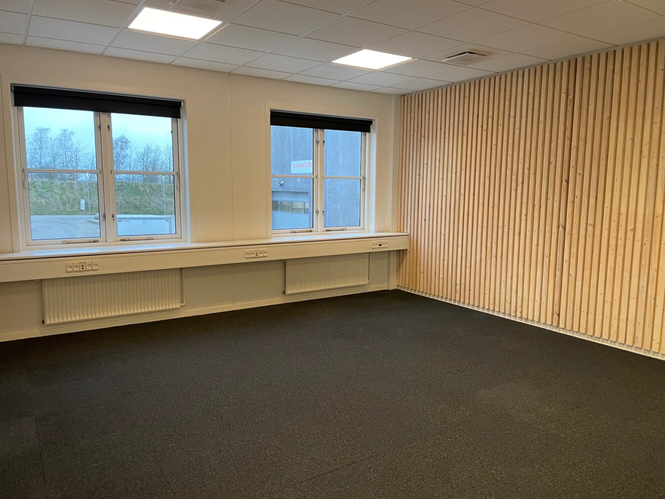 Et tomt kontorlokale med to vinduer, et sort gulvtæppe, hvide vægge og en væg med trælameller.