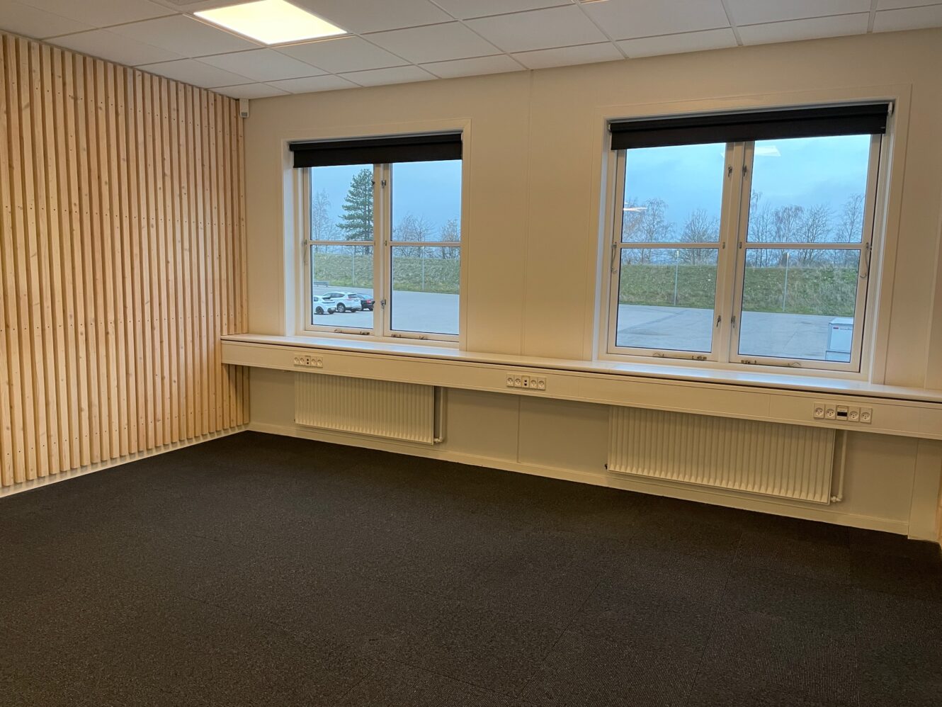 Et tomt rum med sort gulvtæppe, træpanelvæg på den ene side, to store vinduer med sorte persienner og stikkontakter langs væggen under vinduerne.