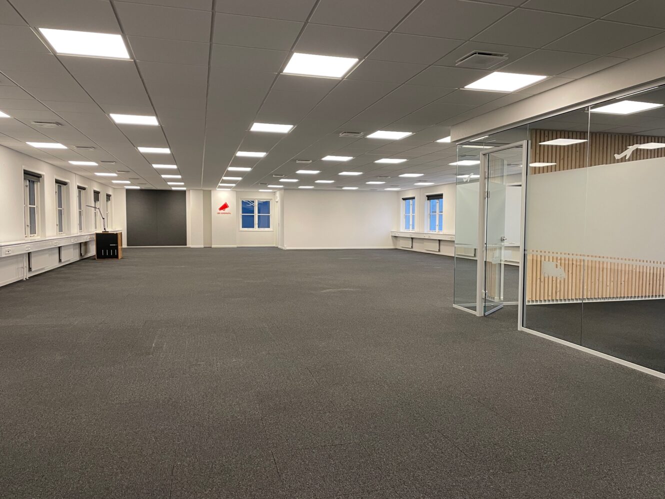 Tomme, moderne kontorlokaler med gulvtæppe, hvide vægge, store vinduer, ovenlys og et mødelokale med glasvægge til højre.