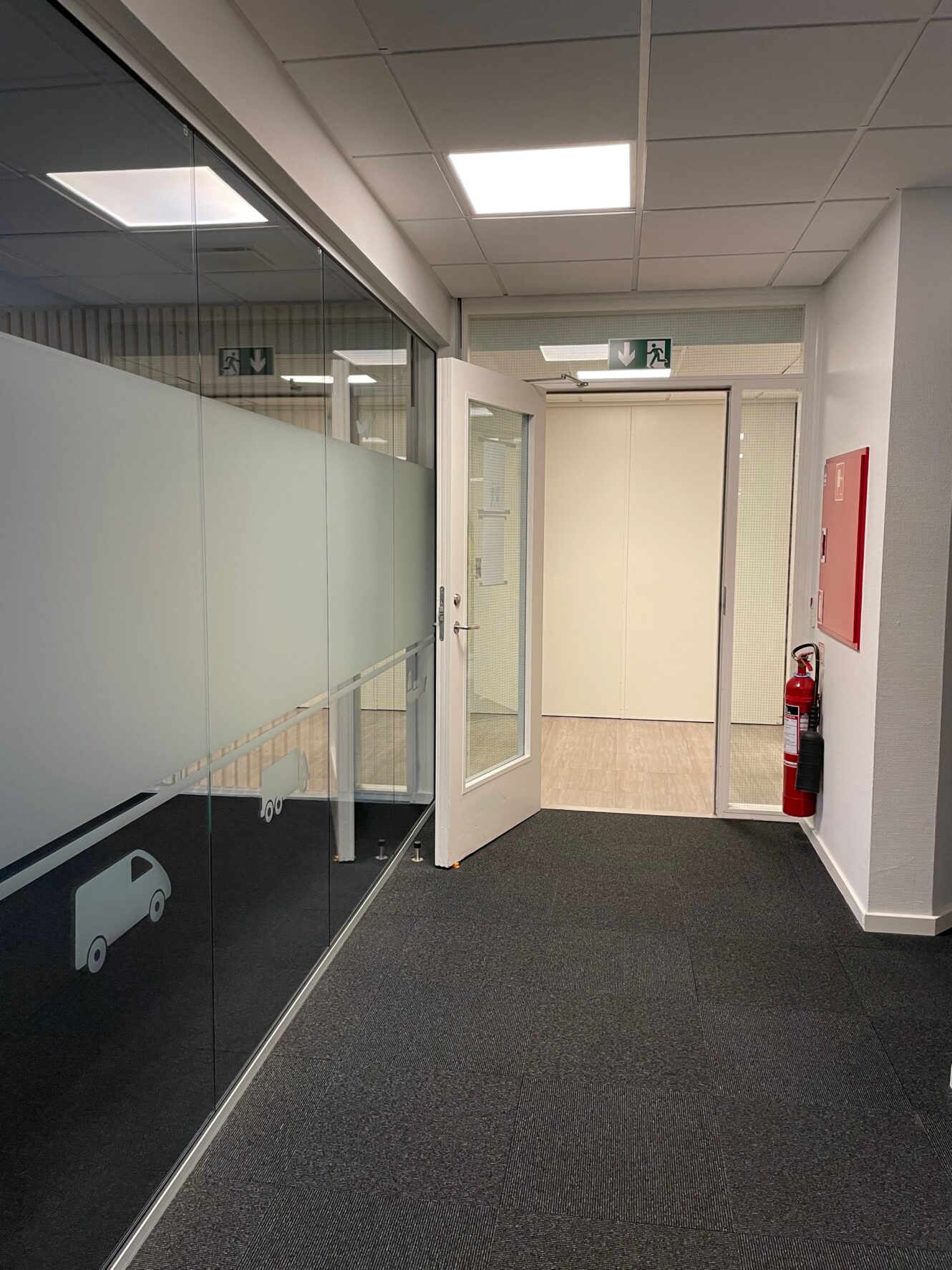 Kontorgang med glasvægge til venstre dekoreret med varebilsmærkater, åben hvid dør for enden, brandslukker og brandalarm på højre væg og ovenlys.