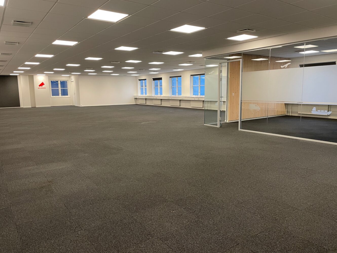 Rummeligt, tomt kontorområde med gråt gulvtæppe, hvide vægge, loftslamper, store vinduer og et mødelokale med glasvægge til højre.