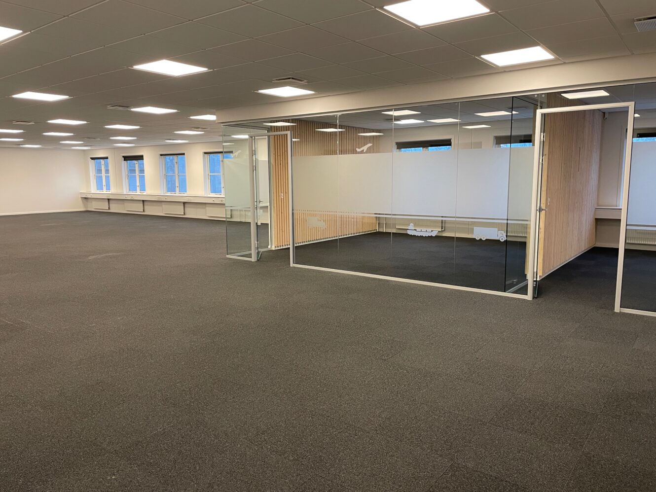 Tomt, moderne kontorlokale med gulvtæppe, store vinduer og et mødelokale med glasvægge. Loftslamperne er tændt, og der er ingen møbler til stede.