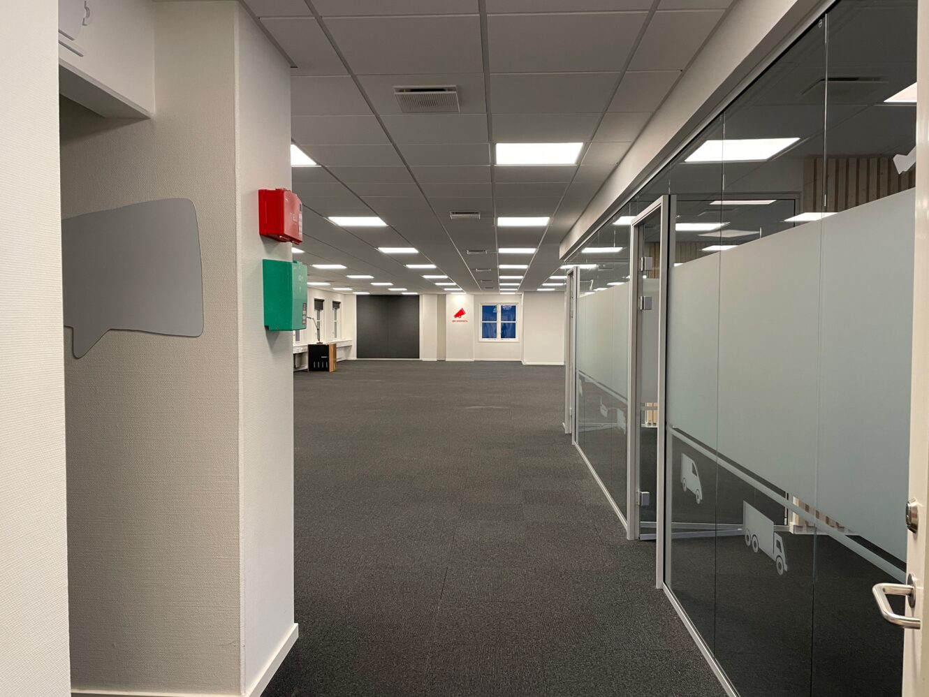 Et moderne kontorlokale med gråt gulvtæppe, mødelokaler med glasvægge til højre og loftslamper, der oplyser et åbent gulvområde.
