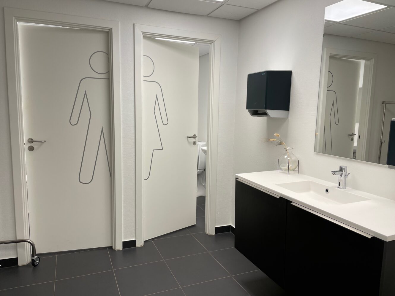 Et moderne toilet med håndvask, spejl, sæbedispenser og to døre markeret med mande- og kvindesymboler.