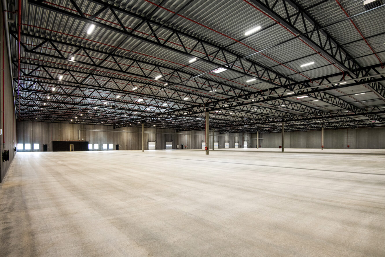 Stort, tomt industrilager med betongulv, metalbjælker og højt til loftet. Lyset kommer ind gennem vinduer langs den fjerneste væg.