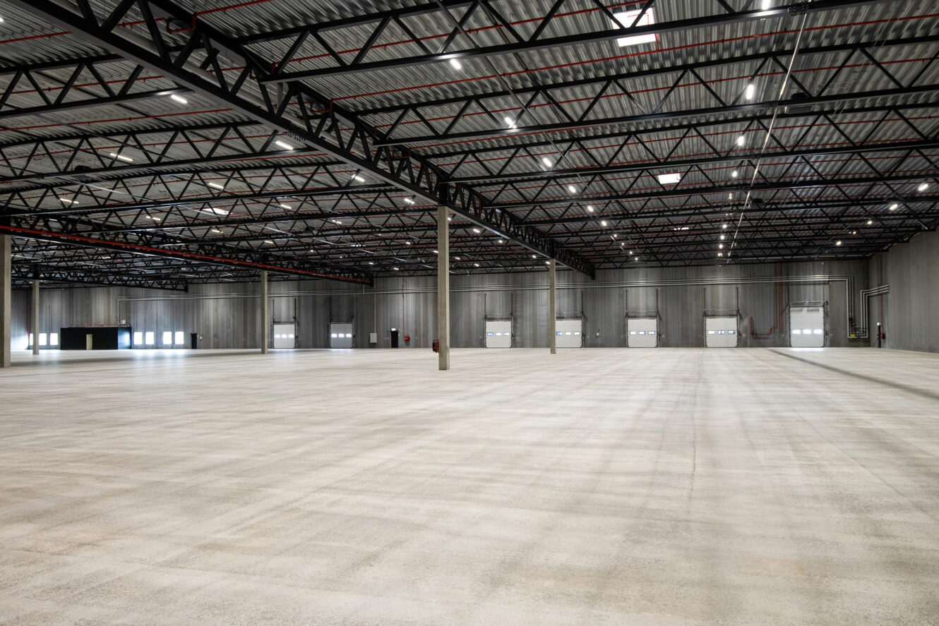 Rummelig tom lagerbygning med højt til loftet, synlige stålbjælker, betongulv og flere læsseramper langs den fjerneste væg.