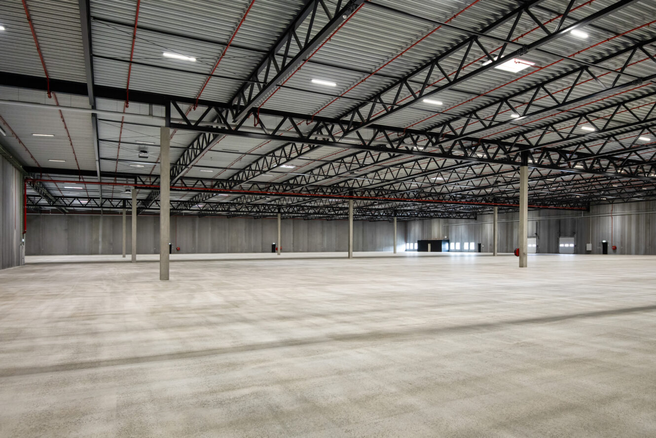 Stort, tomt industrilager med betongulv, metalbjælker og højt til loftet. Lokalet er godt oplyst med ovenlys og naturligt lys fra vinduer.