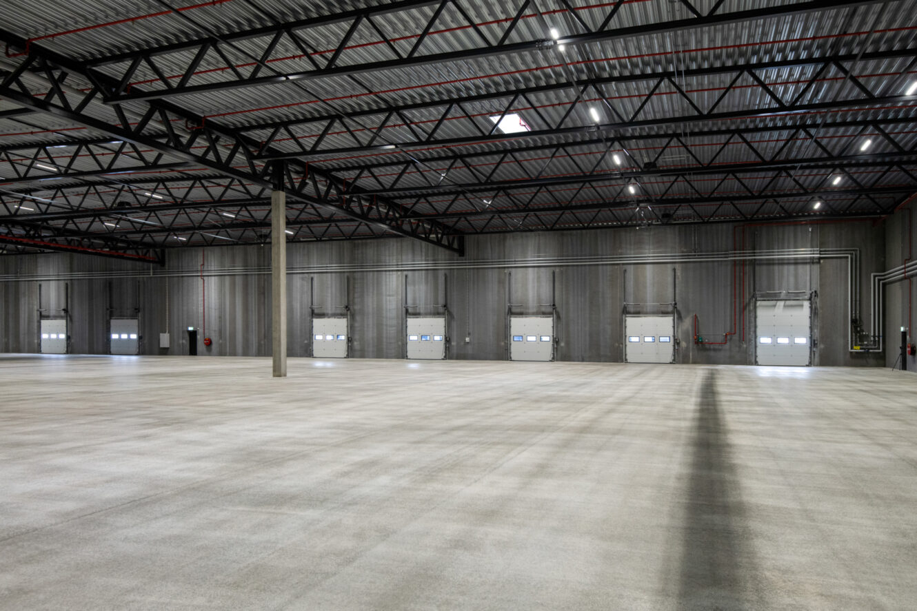 Tomt industrilager med betongulv, grå vægge, synlige stålbjælker og flere porte til læsseramper langs den fjerneste væg.