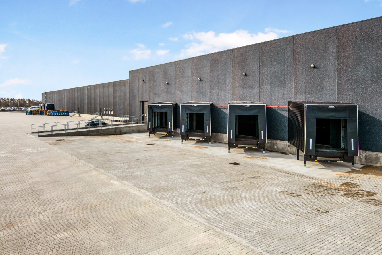 Et stort industrilager med fire læsseramper, hver udstyret med et læskur, der støder op til en stor asfalteret grund under en blå himmel.