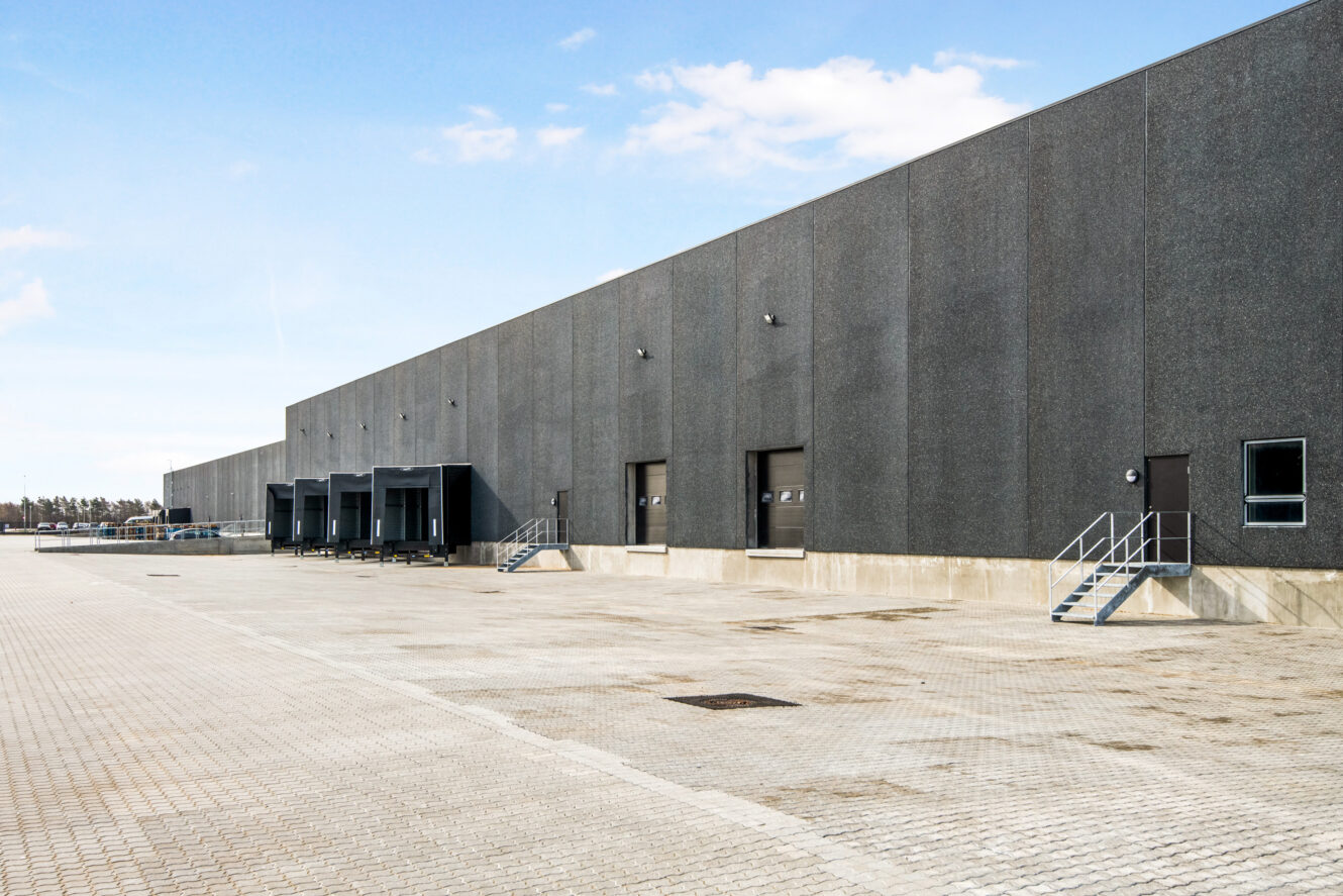 Stort industrilager med flere læsseramper og trapper, beliggende på en asfalteret grund under en delvis overskyet himmel.