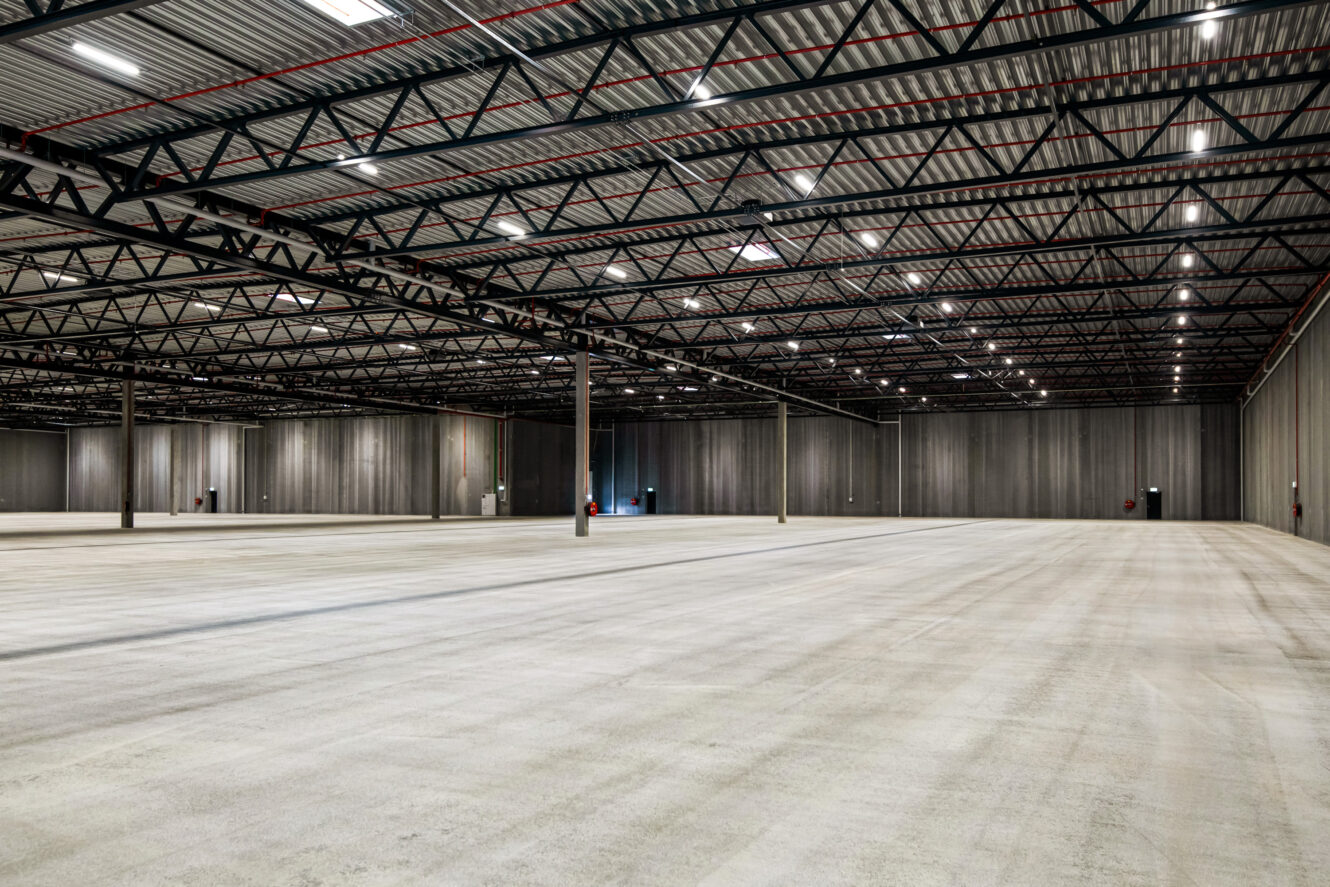 En stor, tom lagerbygning med højt til loftet, synlige bjælker og betongulv. Rummet er godt oplyst med rækker af ovenlys.
