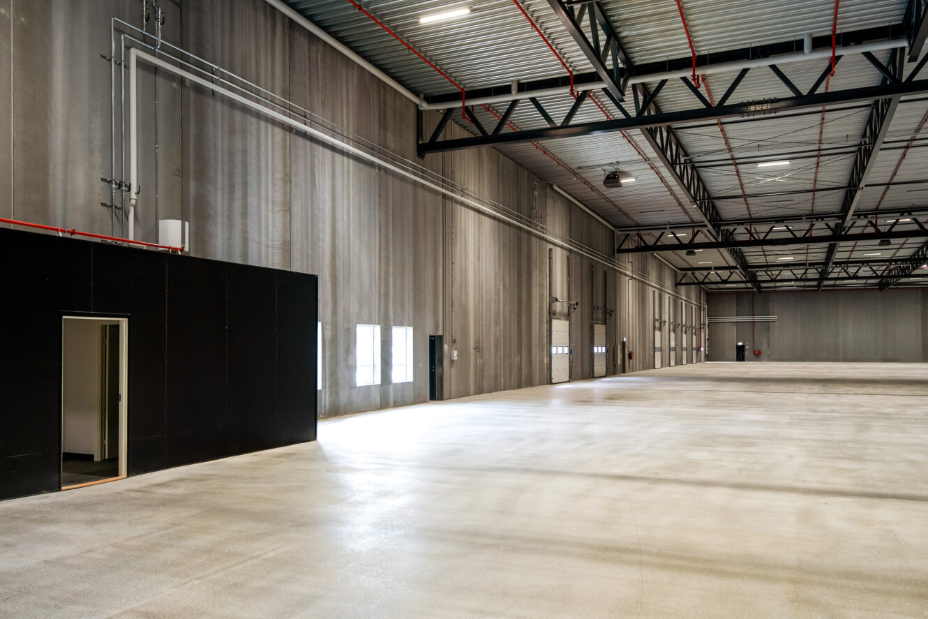 Interiør i en stor tom lagerbygning med betongulv, højt til loftet, synlige bjælker og et lille rum med sorte vægge til venstre.