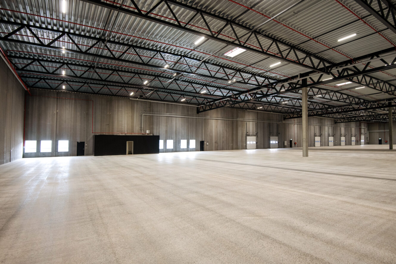 Rummeligt, tomt industrilager med højt til loftet, synlige bjælker, betongulve og store vinduer langs den ene væg.