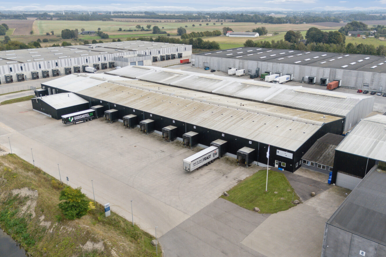 Luftfoto af et stort industrielt lagerkompleks med flere læsseramper og flere lastbiler parkeret udenfor, omgivet af lignende lagerbygninger og åbne marker.