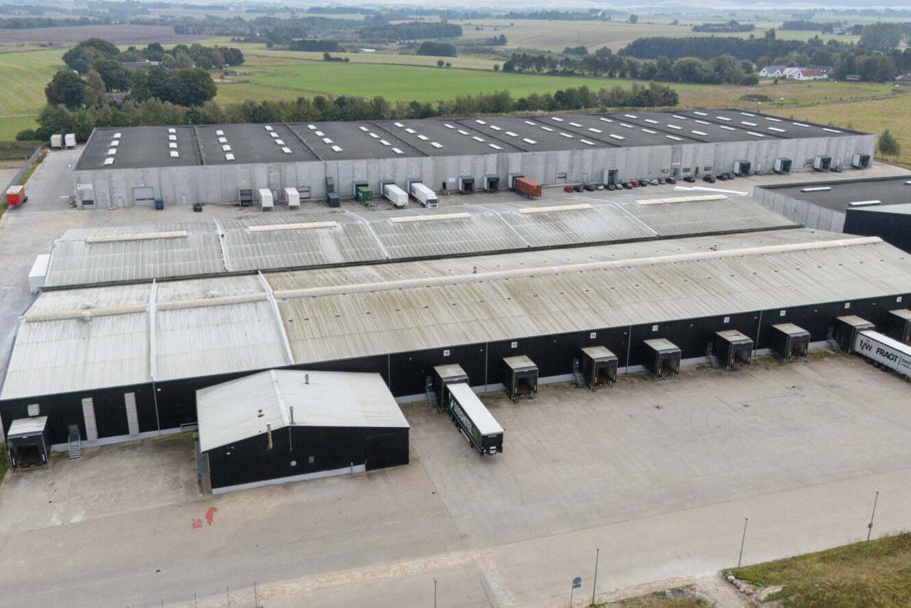 Luftfoto af et stort industrielt lagerkompleks med flere læsseramper og parkerede lastbiler, omgivet af marker og landskab.