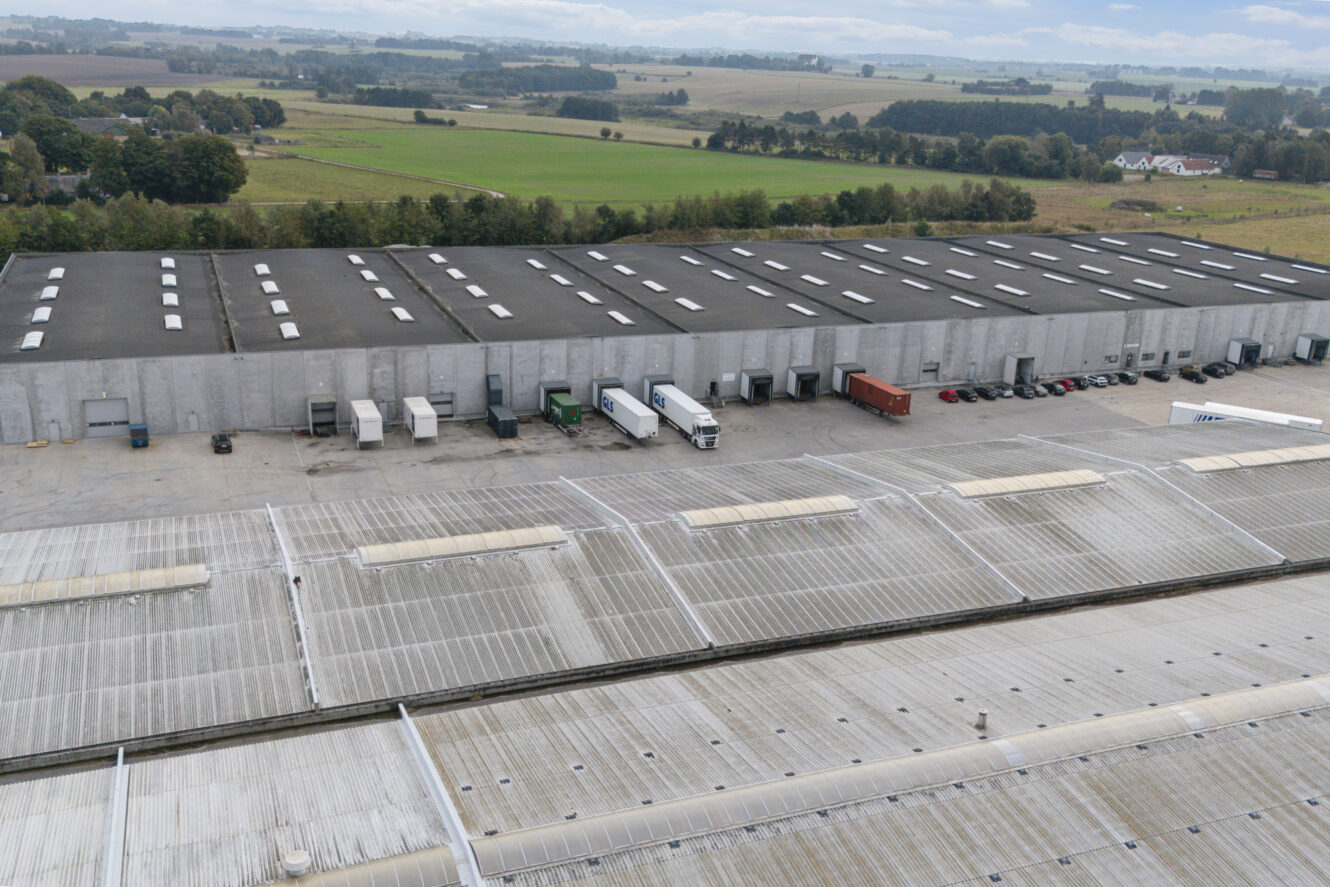 Luftfoto af et stort industrilager med læsseramper, lastbiler, parkerede biler og åbne marker i baggrunden.