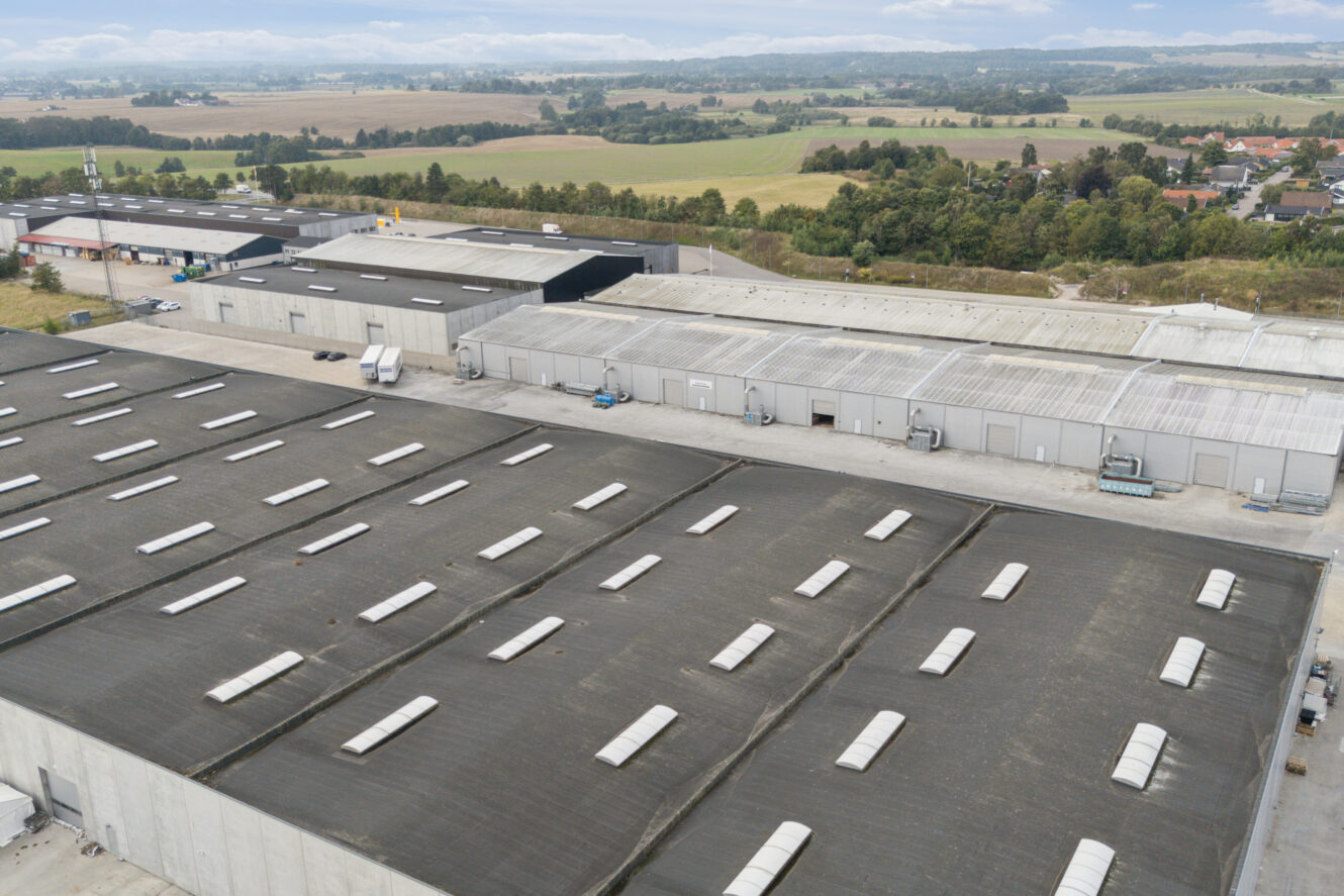 Luftfoto af store industrilagre med mørke tage, omgivet af veje, marker og spredte træer på en overskyet dag.