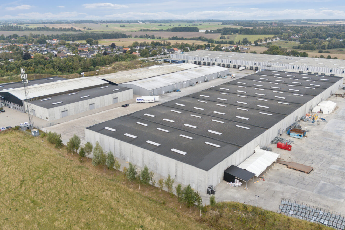 Luftfoto af store industrielle lagerbygninger med læsseramper, omgivet af asfalterede områder og marker, under en delvist overskyet himmel.
