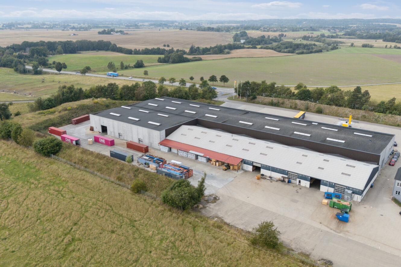 Luftfoto af et stort industrilager med læsseramper, skibscontainere og lastbiler, omgivet af marker og fjerne træer under en delvis overskyet himmel.