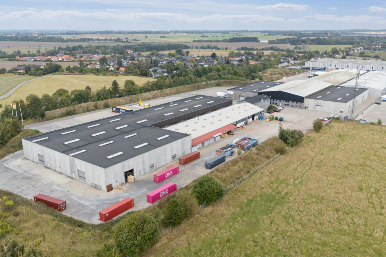 Luftfoto af et industrielt lagerkompleks med skibscontainere og åbne gårdspladser, omgivet af marker og et fjernt boligområde.