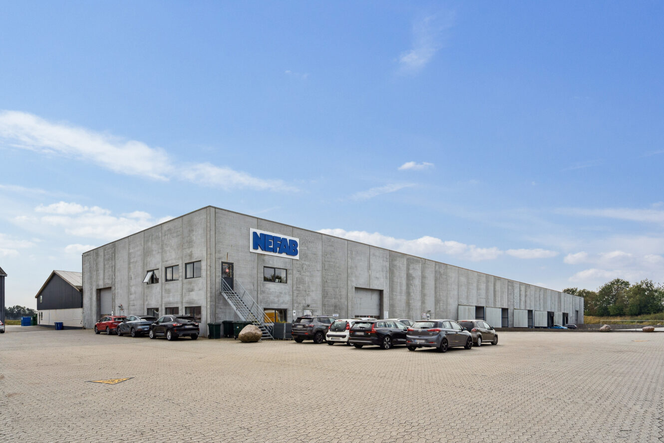 Et stort, gråt industrilager med et NERGBS-skilt, flere garageporte og flere biler parkeret foran under en klar blå himmel.