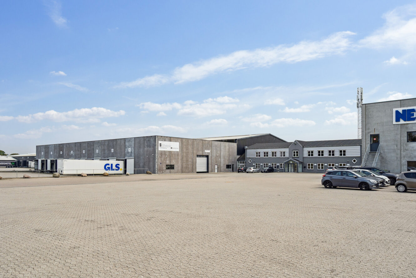 Store industribygninger med en GLS-lastbil, flere parkerede biler og en stor asfalteret plads under en delvis overskyet himmel.