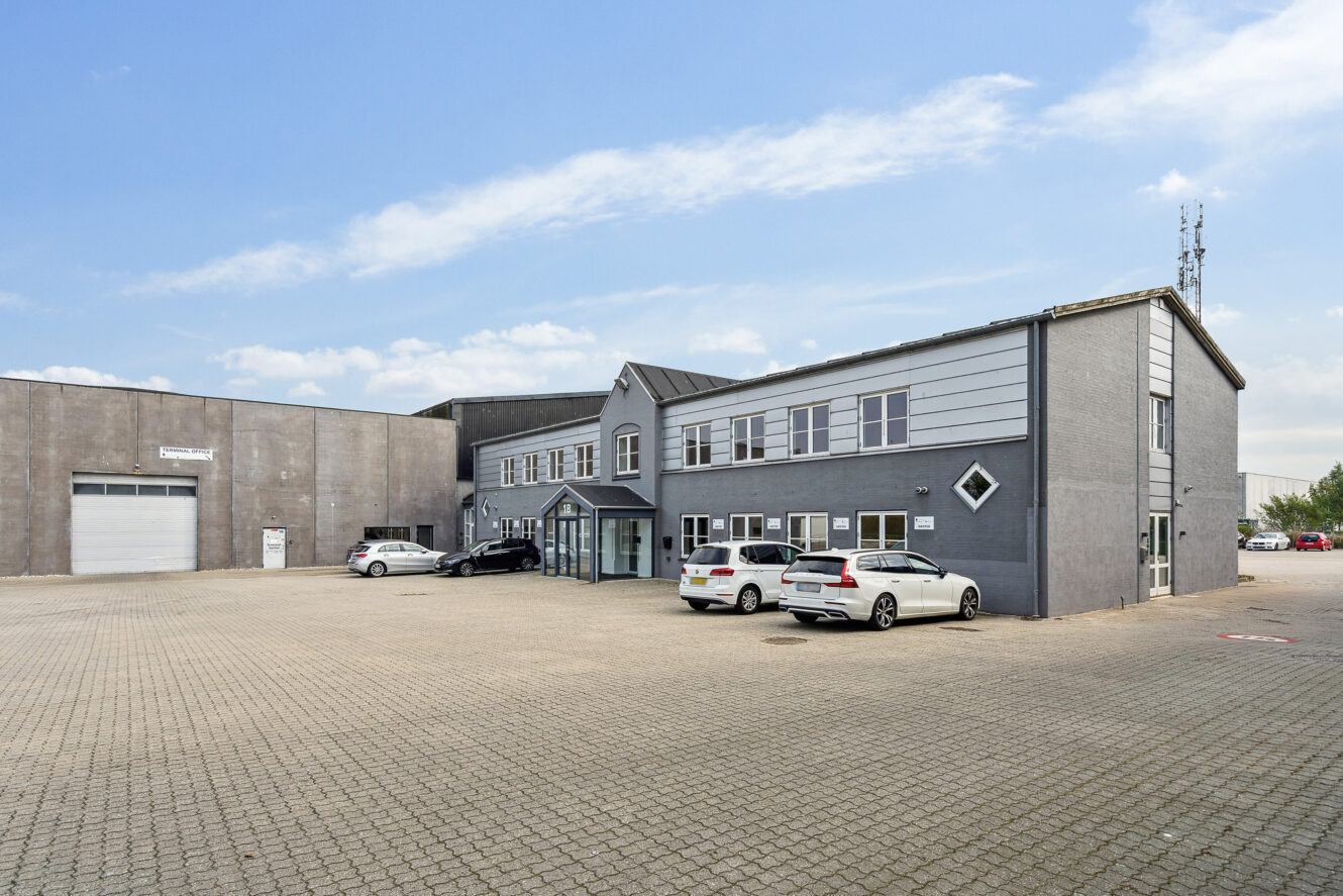 En toetagers grå kontorbygning med flere parkerede biler foran, placeret på en stor asfalteret grund under en delvist overskyet himmel.