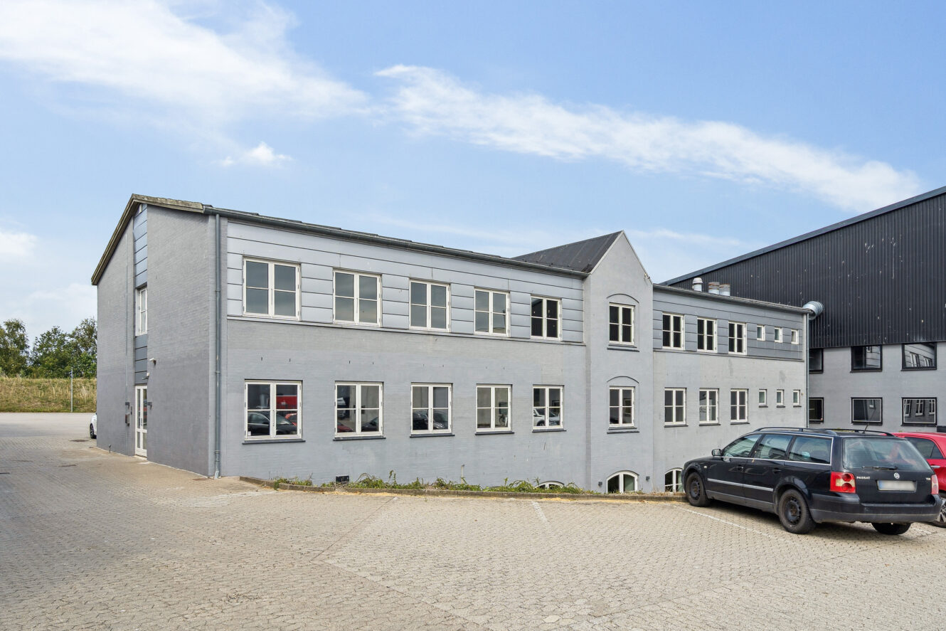 En toetagers grå bygning med flere vinduer og en spids indgang, der støder op til en sort bygning, med biler parkeret foran på en asfalteret grund.