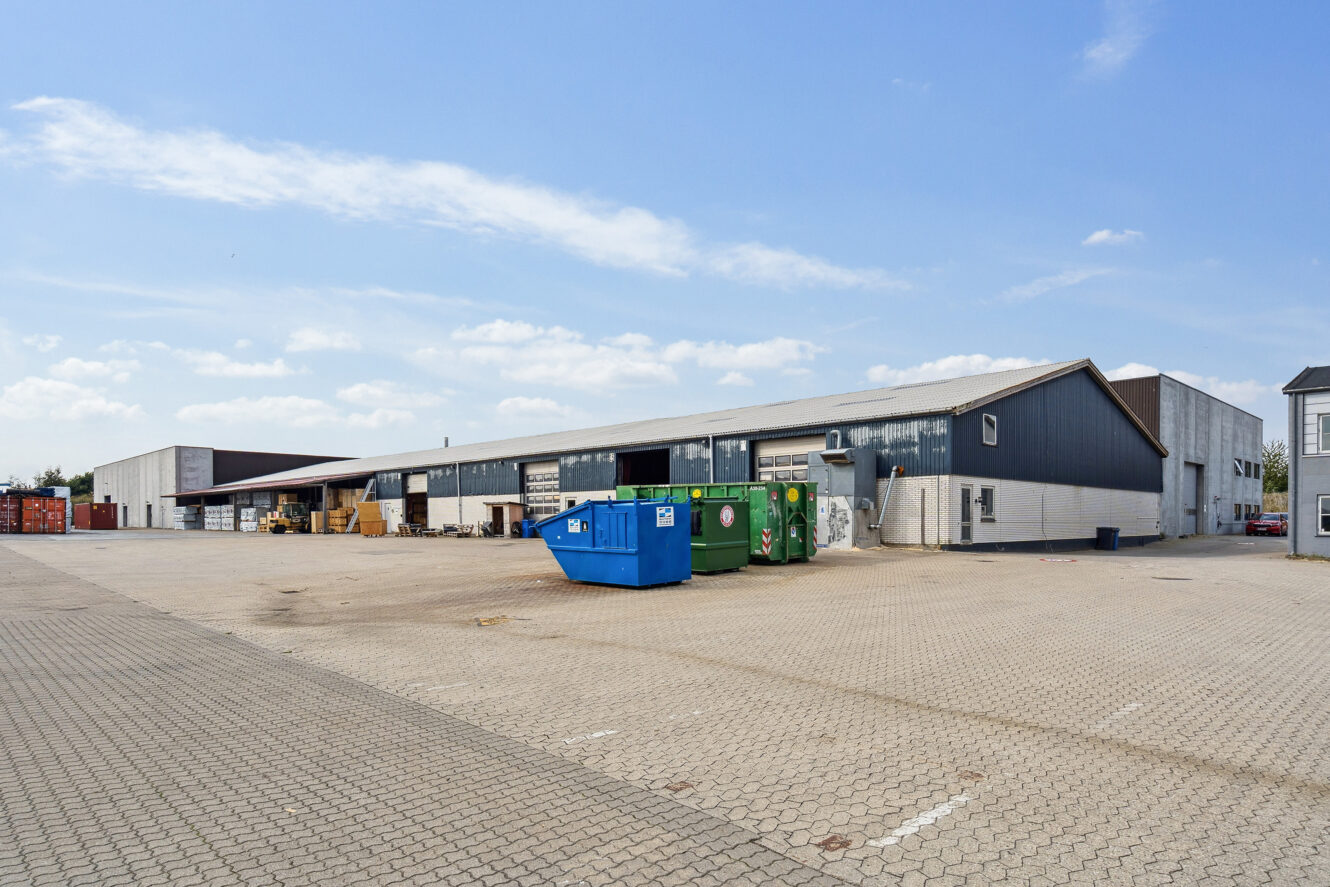 Et stort industrilager med en asfalteret plads, flere containere og stablede paller under en delvis overskyet himmel.