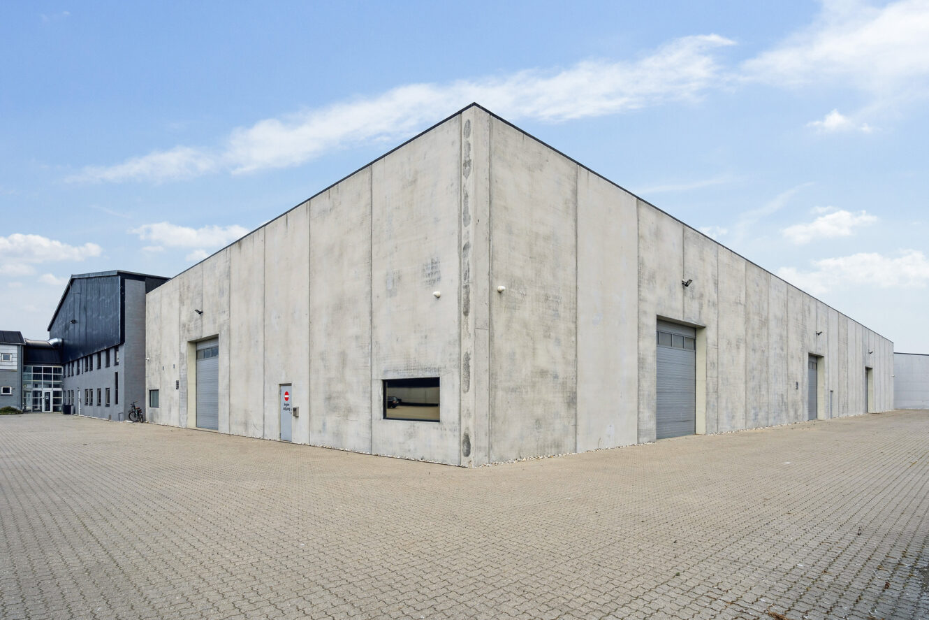 En stor, moderne lagerbygning med grå betonvægge og flere lukkede rulleporte ligger på en asfalteret grund under en delvis overskyet himmel.